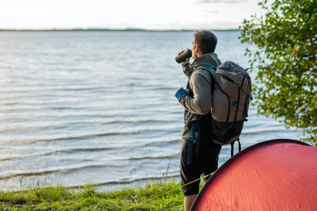 Man camping in Estonia, standing at lake with a backpack, drinking coffee